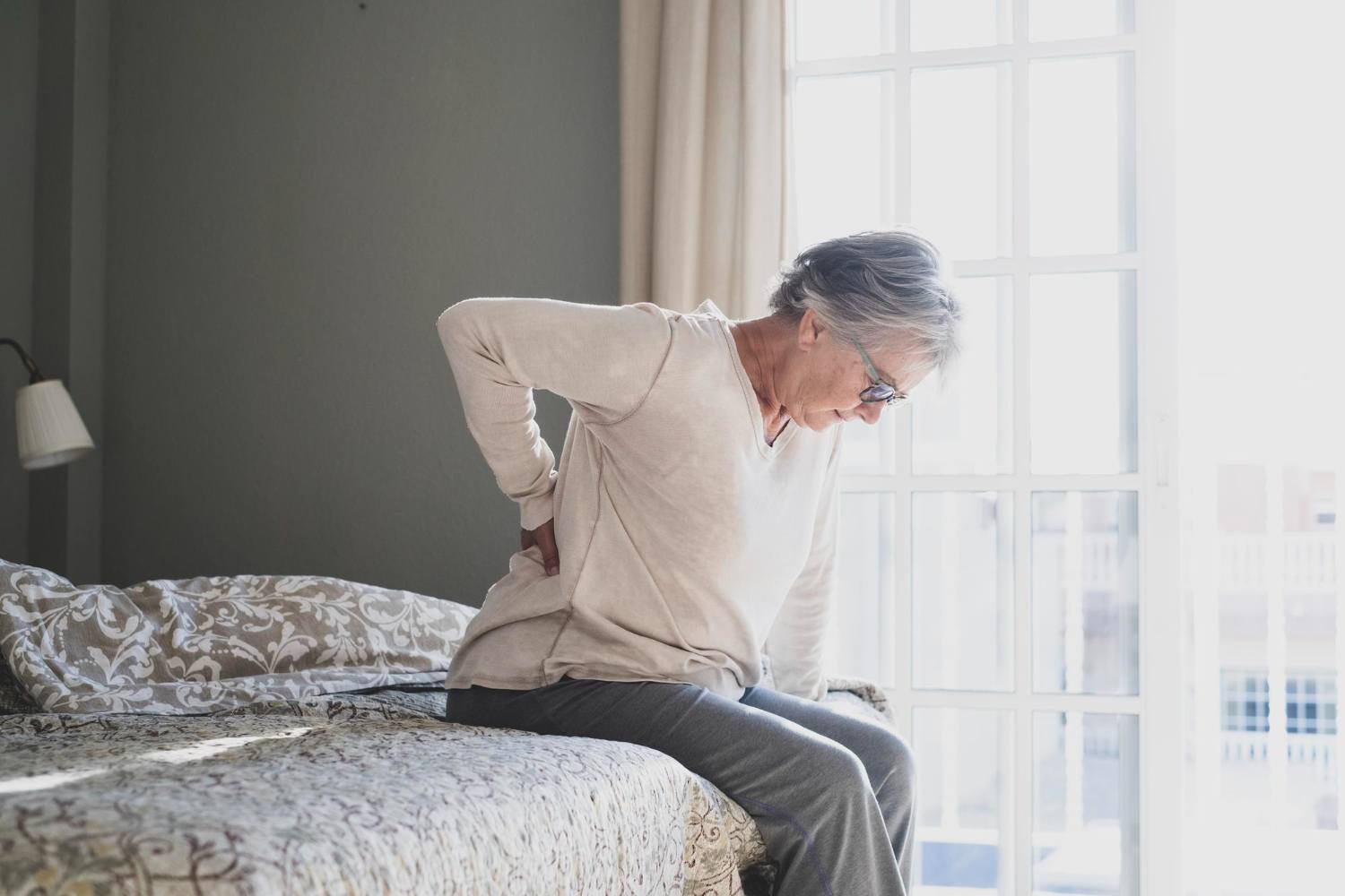 Older woman sitting on her bed and holding her lower back, a common sign of spinal pain.